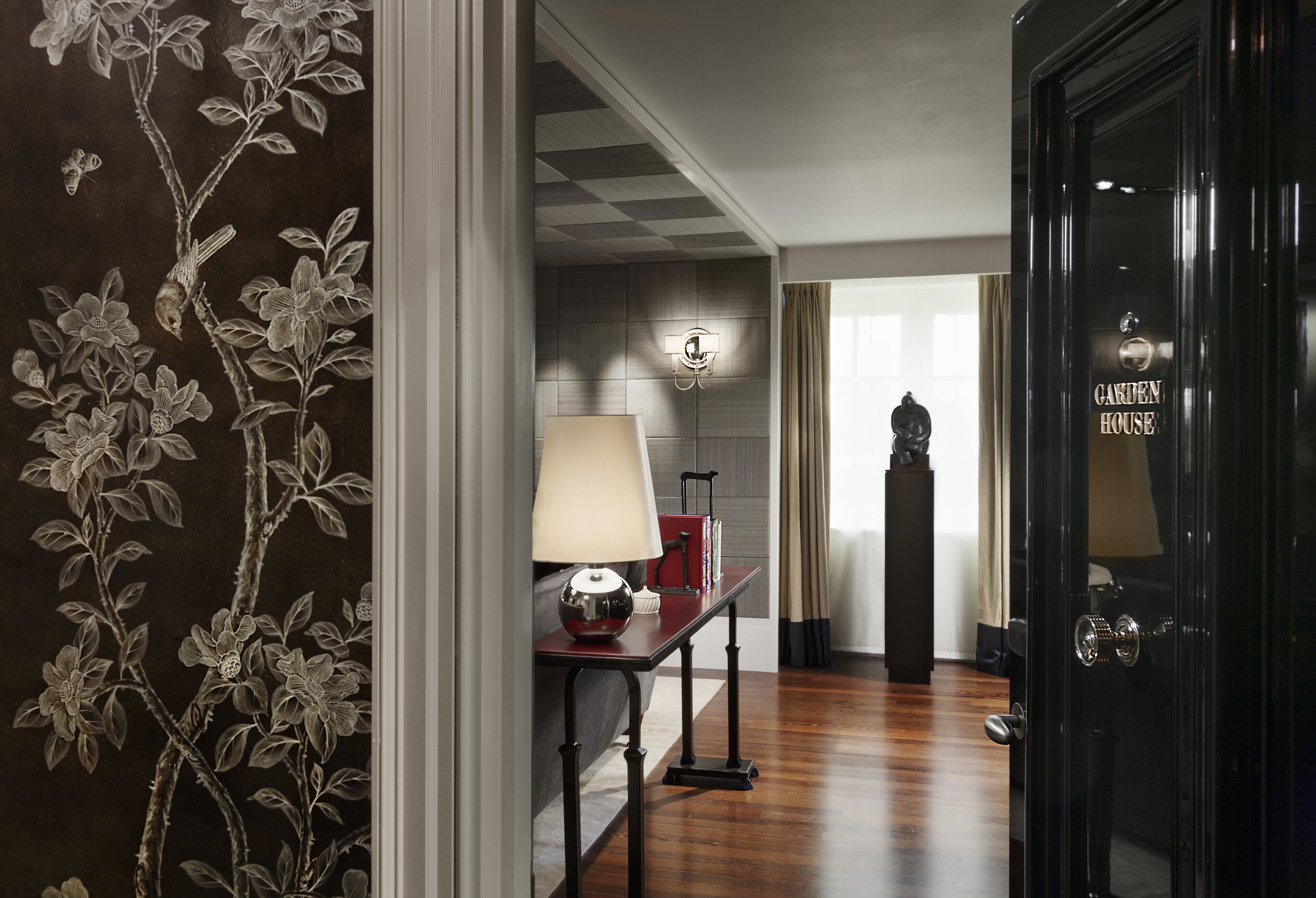 A hallway with a dark wooden door labeled "GARDEN HOUSE," a lamp on a table, and a wall with a floral pattern.