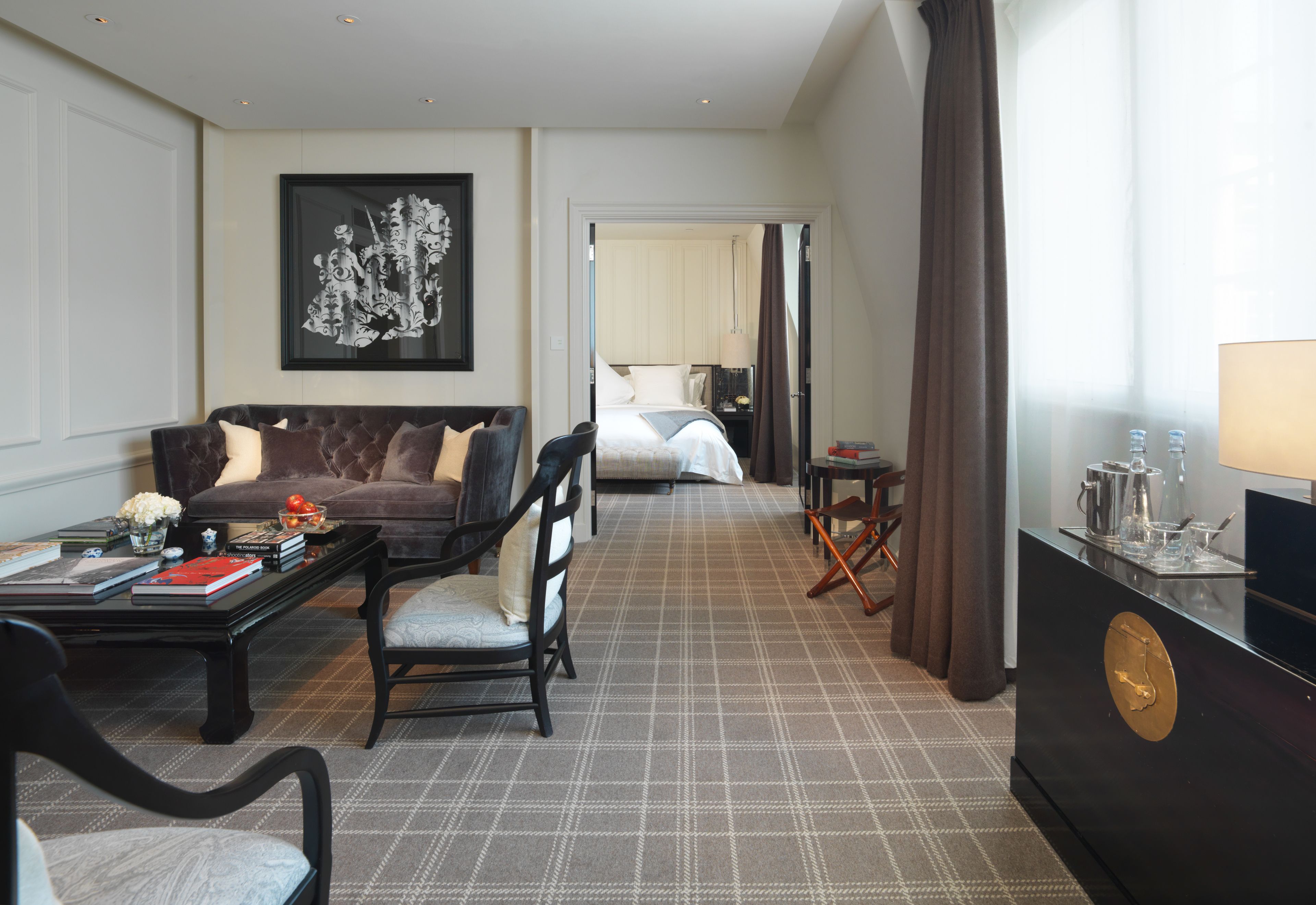 Habitación de hotel con sofá, sillón y cama. Hay un escritorio con libros y un florero. Un cuadro adorna la pared, y una lámpara está sobre la mesita de noche.
