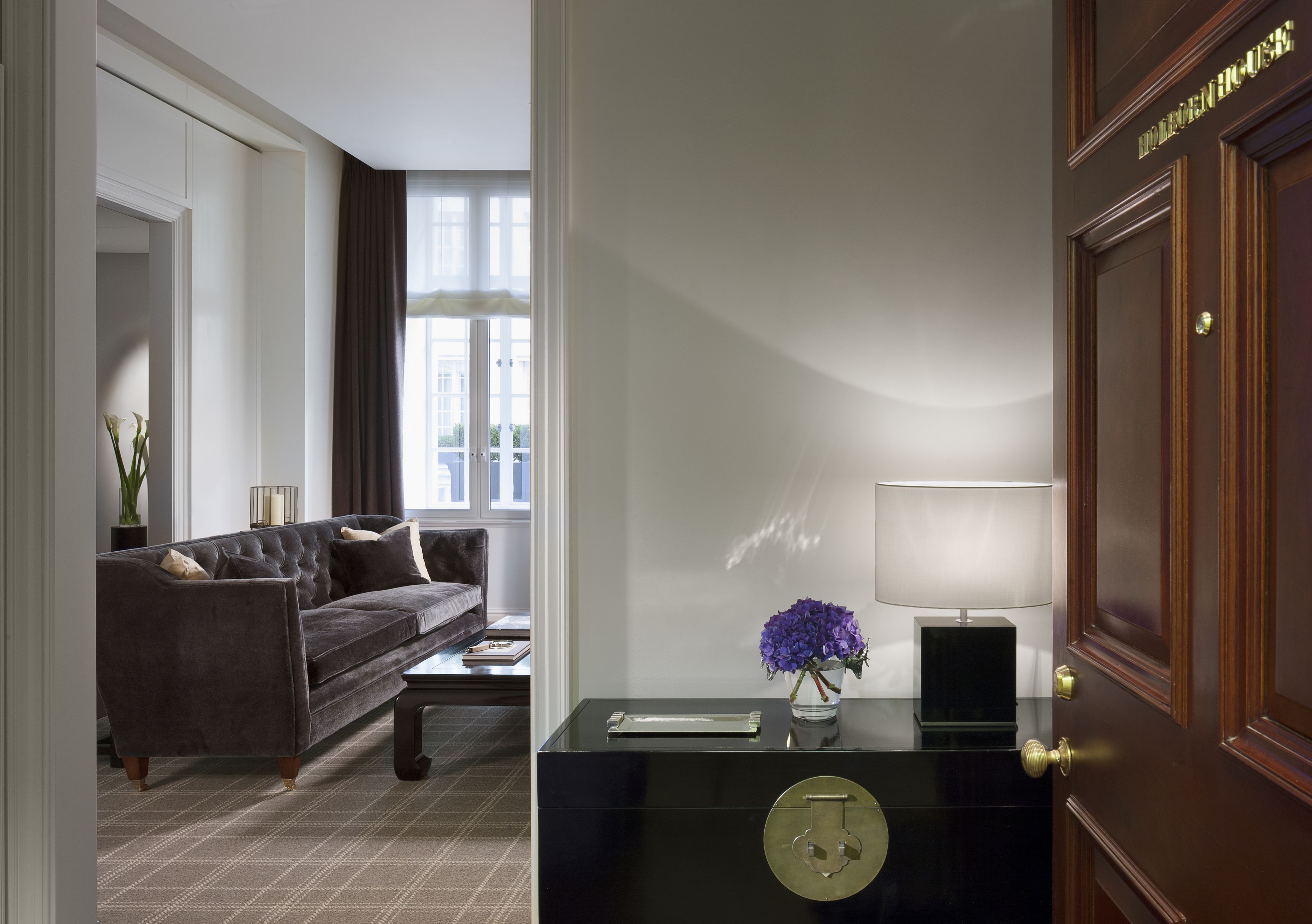 Una habitación de hotel moderna con un sofá de color oscuro, una mesa de centro de vidrio, un escritorio con una lámpara y una puerta con manija de latón.