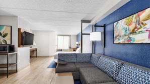 Una moderna camera d'hotel con un divano componibile, una TV a schermo piatto, una scrivania e un grande quadro astratto appeso al muro.