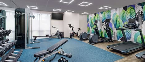 A gym with exercise equipment, a bench, dumbbells, treadmills, and a wall with a leaf pattern.