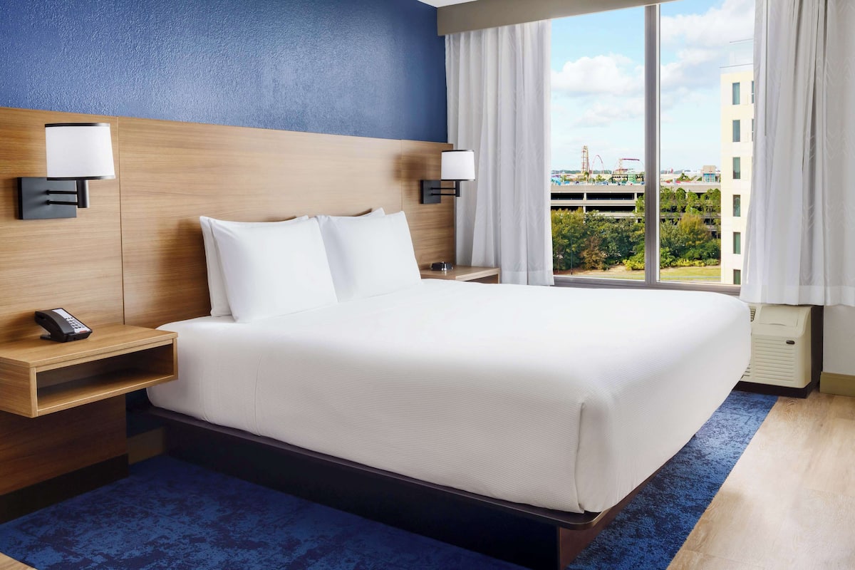 Een hotelkamer met een groot bed, twee nachtkastjes met lampen, een telefoon en uitzicht op de stad door grote ramen.