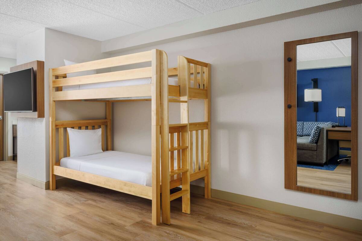 Een stapelbed met houten frame en witte beddengoed, een flatscreen televisie aan de muur bevestigd, en een spiegel die een kamer reflecteert met een blauwe wand en een bureau met een lamp.