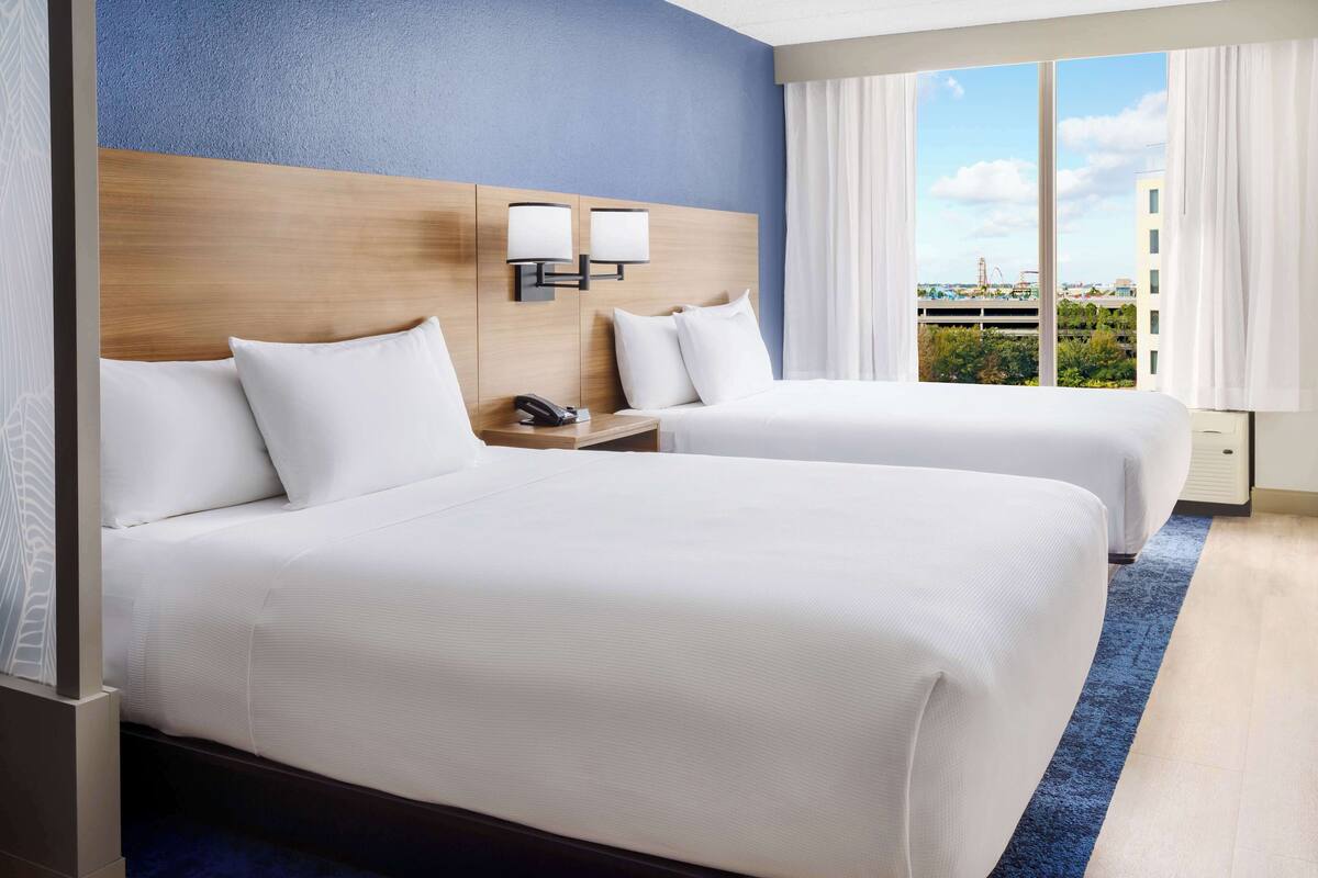 Hotelkamer met twee bedden, een houten hoofdbord, een nachtkastje met telefoon en uitzicht op de stad door grote ramen.