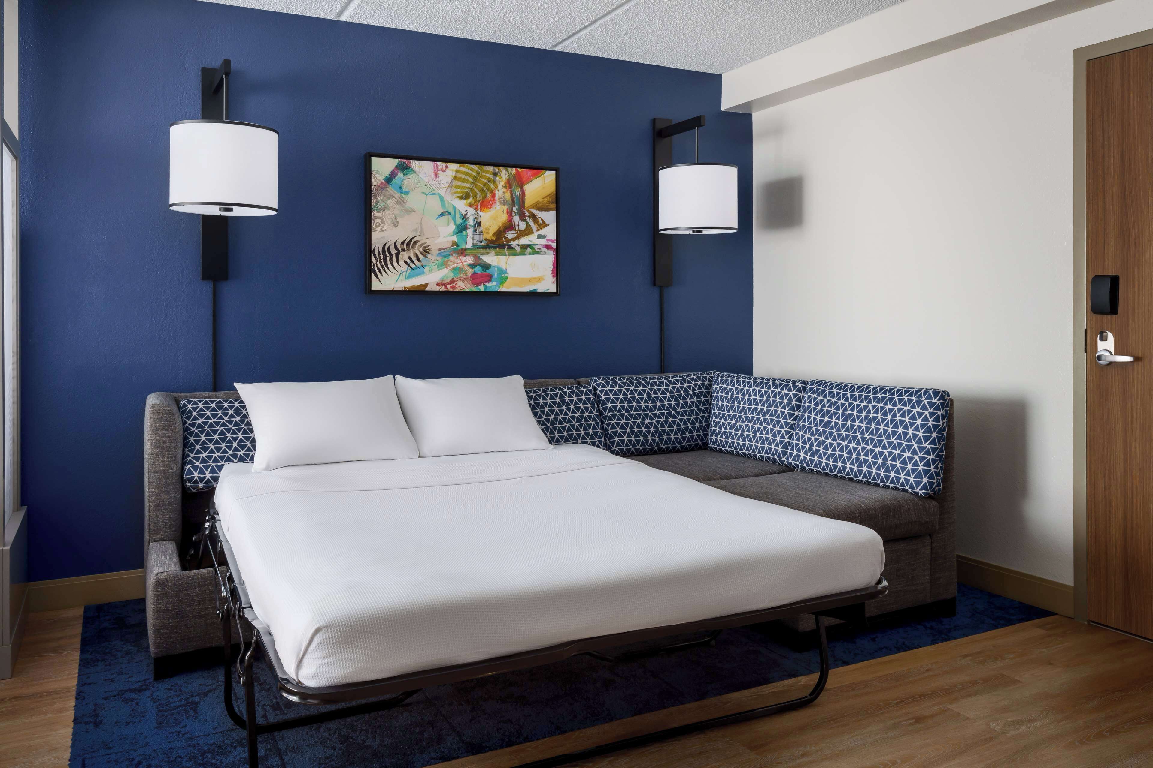 Una habitación de hotel moderna con una cama grande, un sofá, dos lámparas de pared y un cuadro abstracto colorido en la pared.