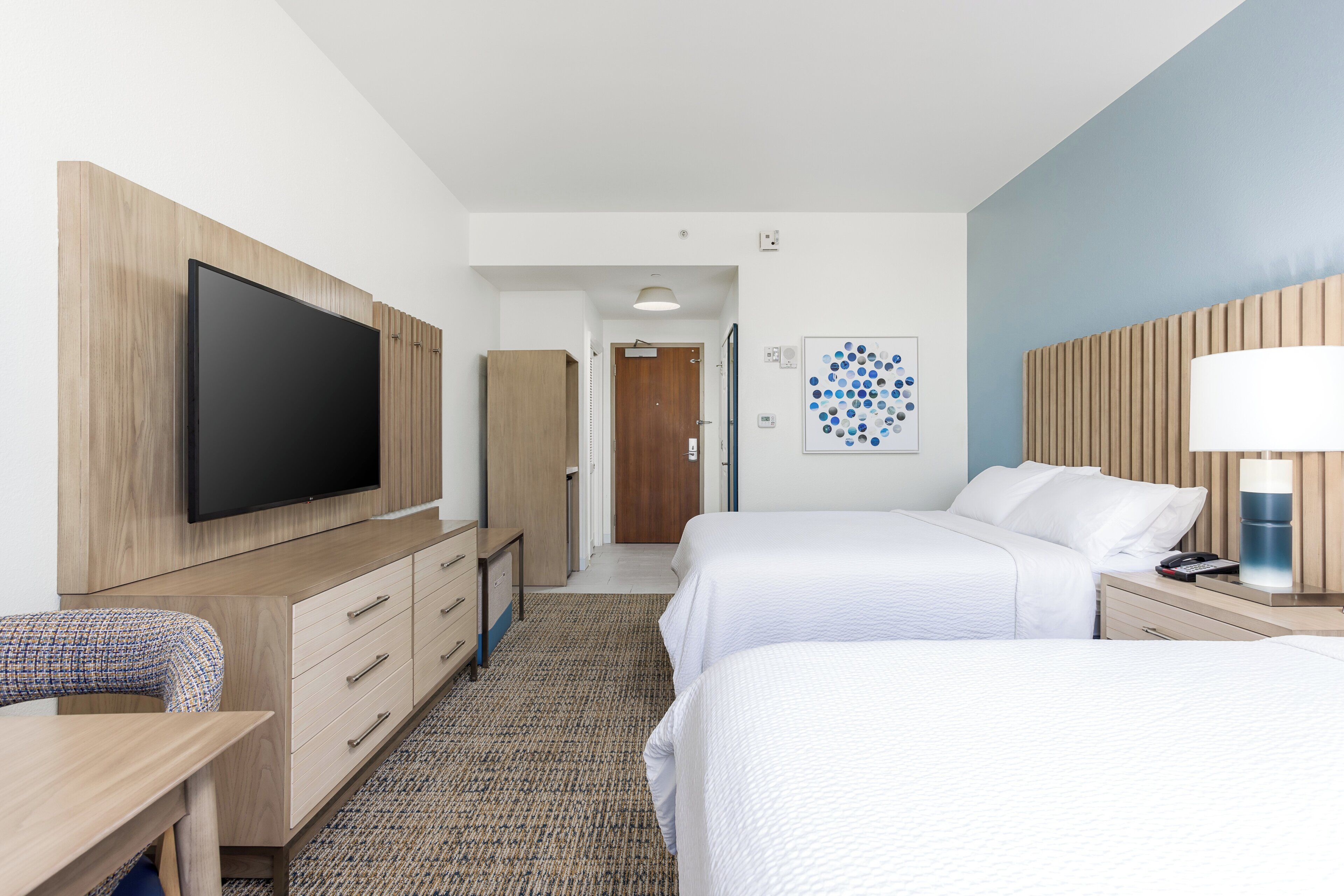 A hotel room with a flat-screen TV, two beds, a desk, and a wall-mounted lamp.