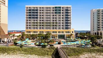 A hotel with a large central building, a pool area with umbrellas, and a beach in the foreground.