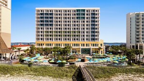 A hotel with a large central building, a pool area with umbrellas, and a beach in the foreground.