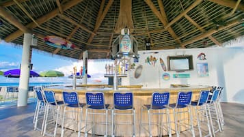 Un bar en la playa con techo de paja, sillas azules y vista al océano.