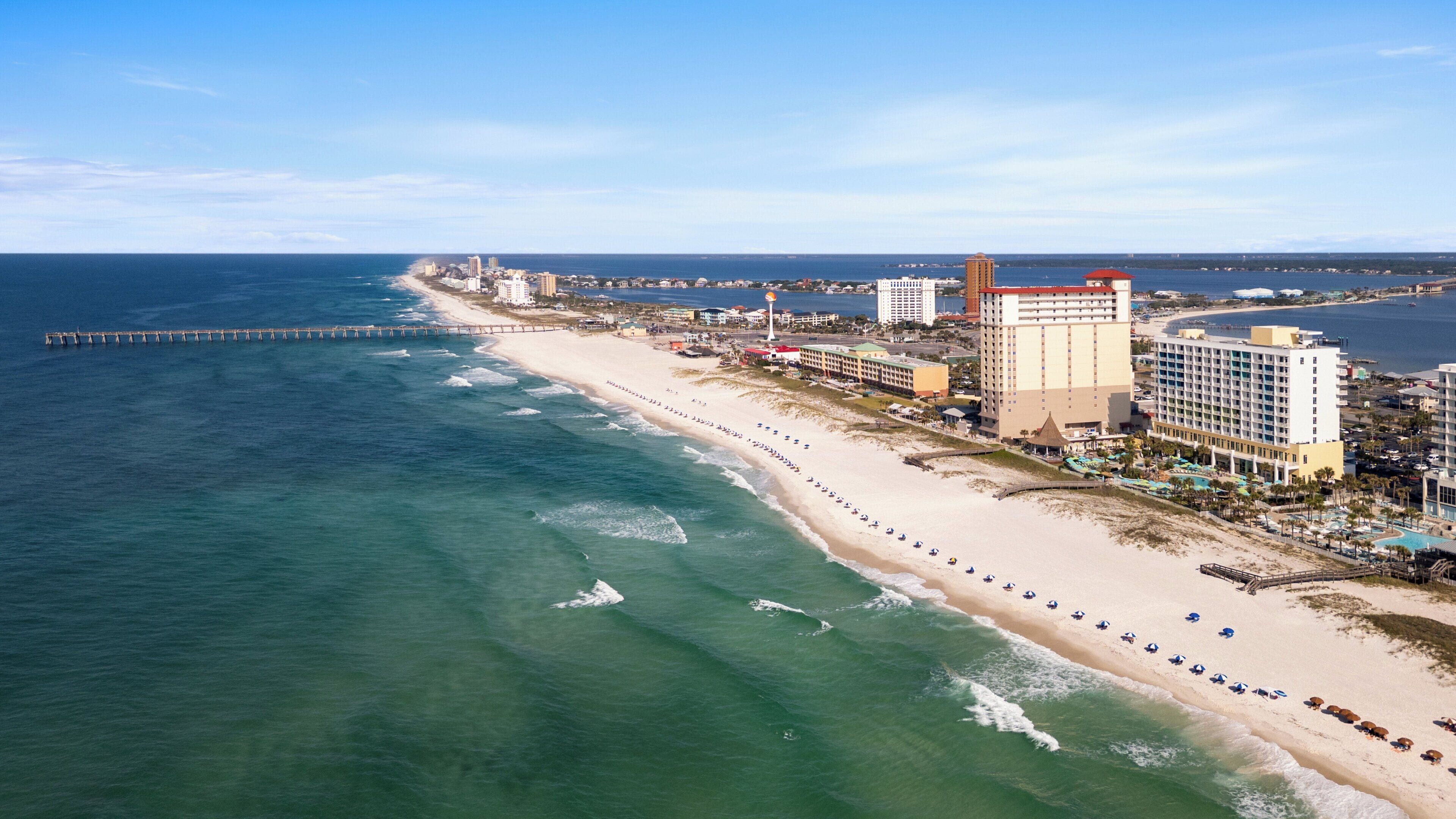 A coastal hotel with a beachfront view, a pier extending into the ocean, and a row of beach umbrellas along the shore.