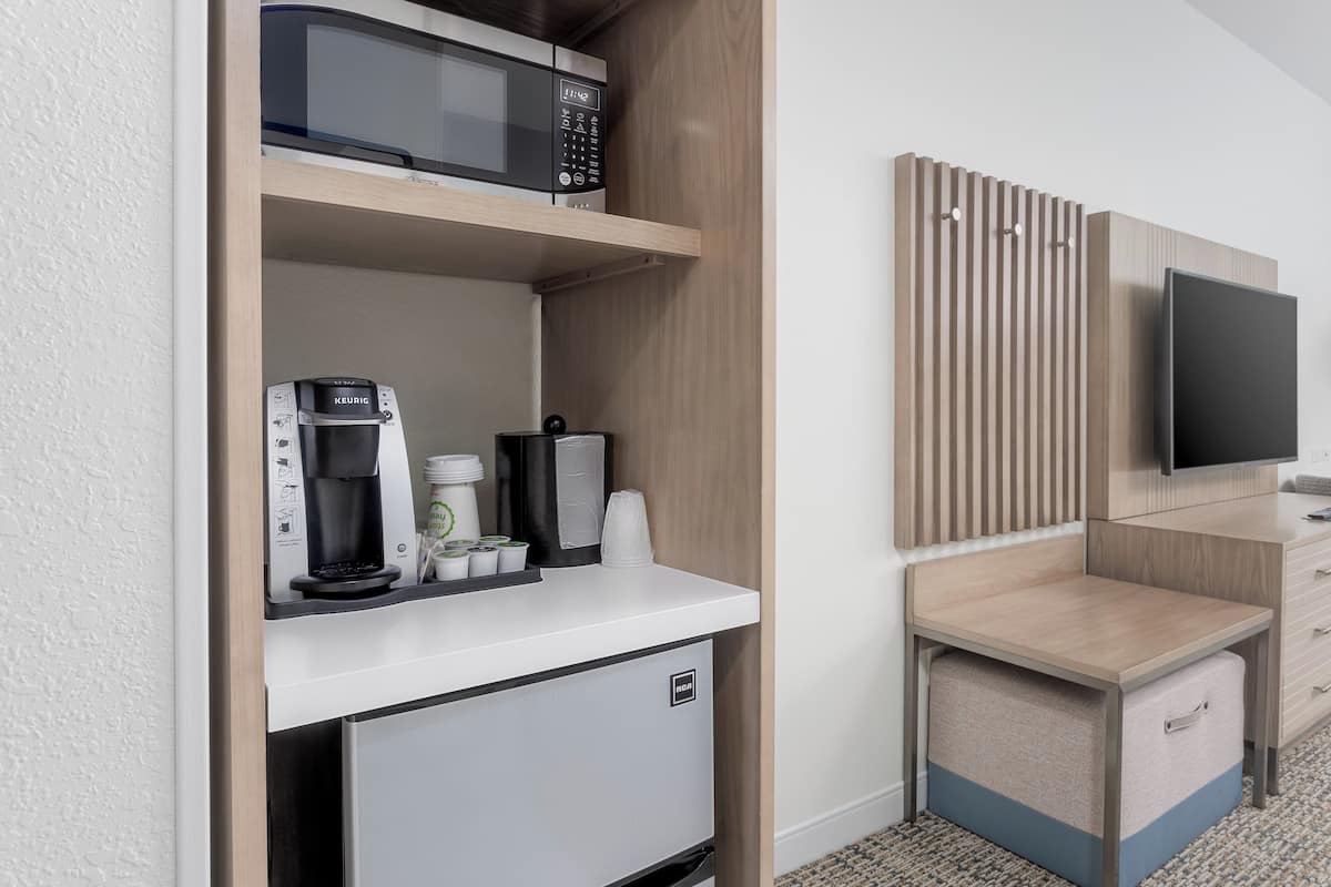 Eine Küchenzeile mit Mikrowelle, Kaffeemaschine und kleinem Kühlschrank. Ein Fernseher ist an der Wand montiert und es gibt eine kleine Sitzecke mit Tisch und Stauraum.
