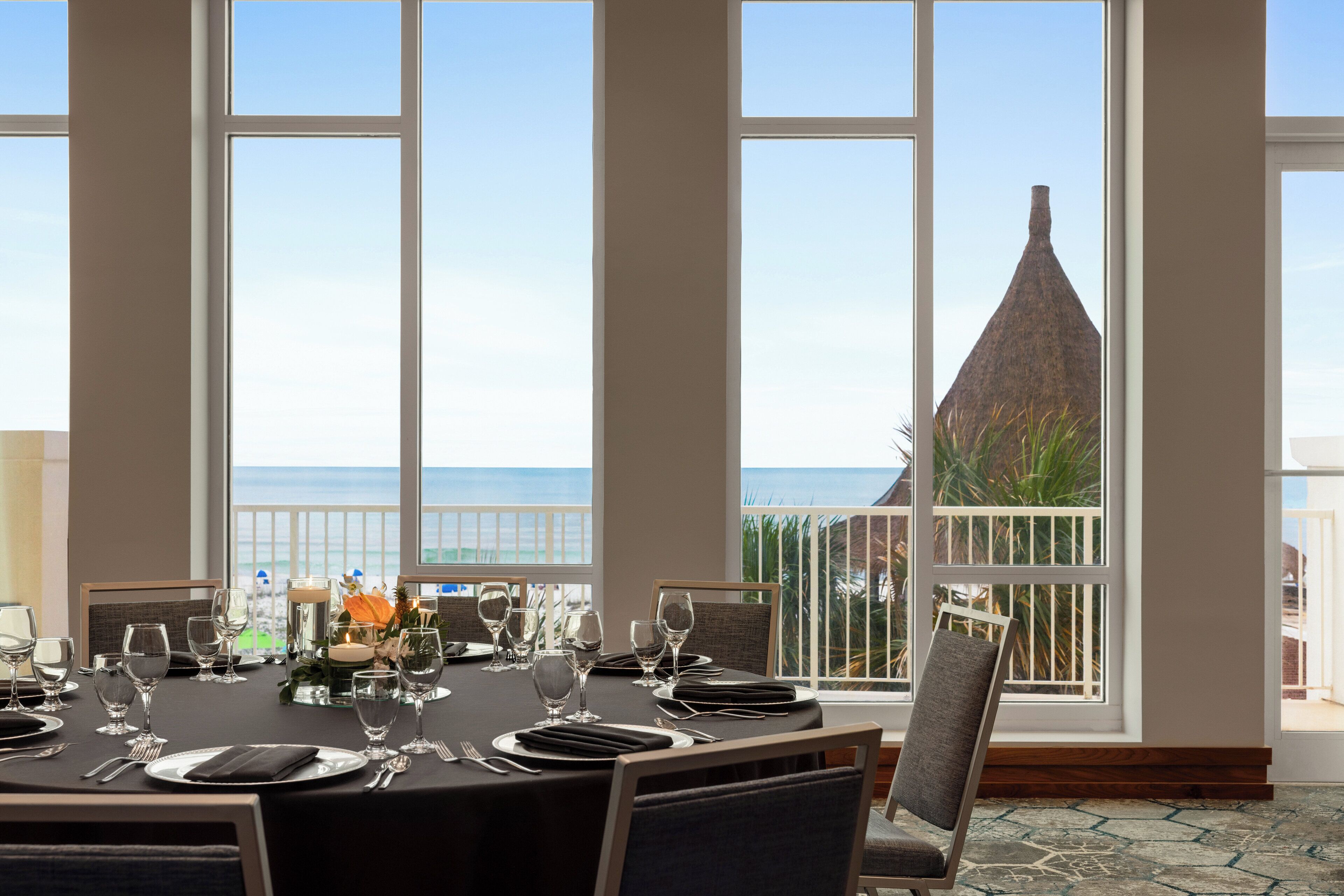 Un comedor con una mesa dispuesta para cuatro personas, ventanas amplias con vista al mar y un edificio con techo cónico.