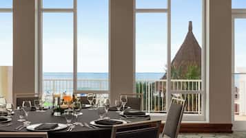 Un comedor con una mesa dispuesta para cuatro personas, ventanas amplias con vista al mar y un edificio con techo cónico.