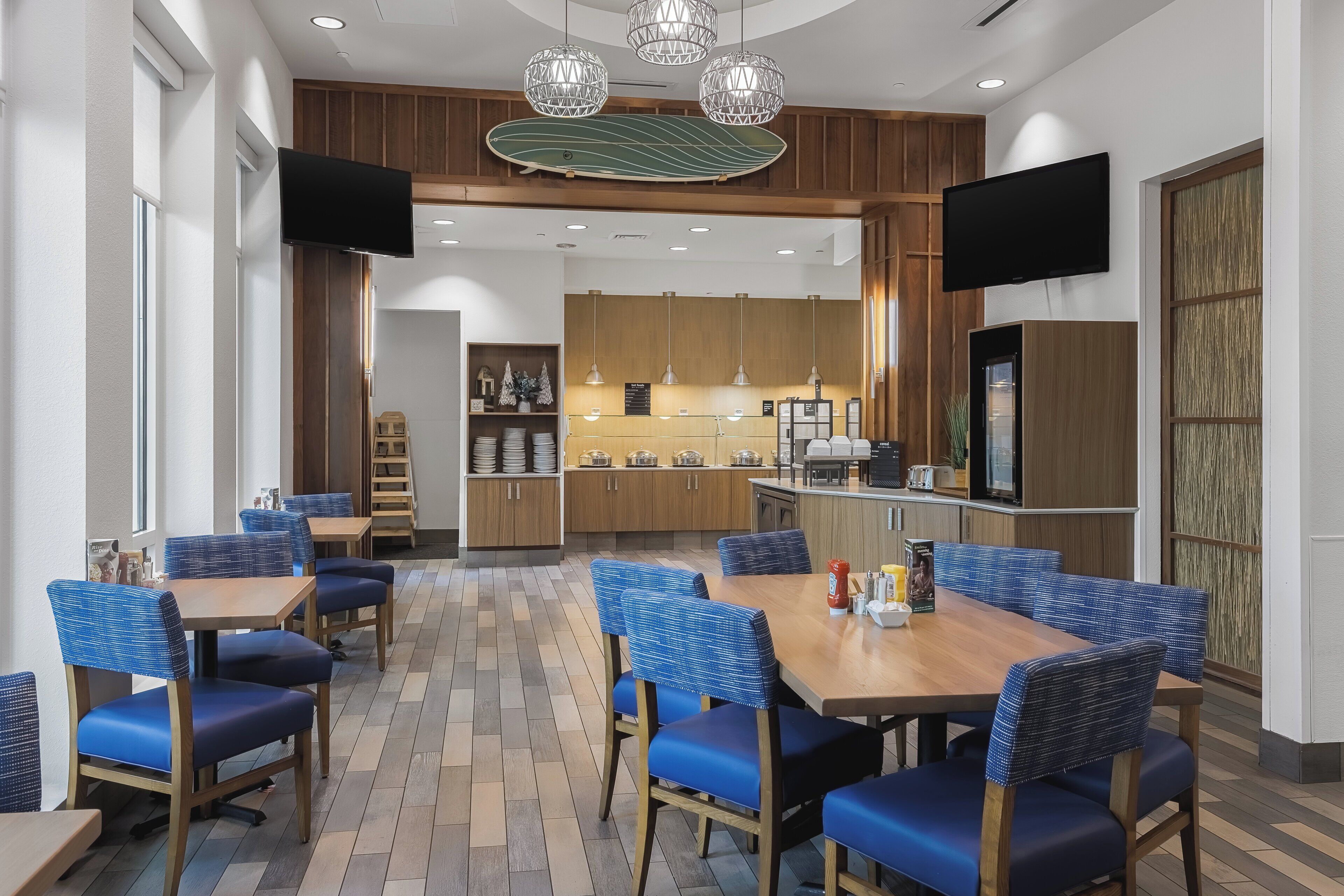 Un espace de salle à manger moderne avec des chaises à revêtement bleu et une table en bois, une cuisine en arrière-plan et un éclairage suspendu.