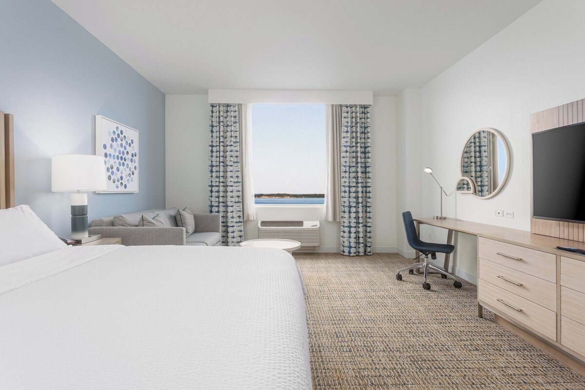 A hotel room with a bed, a desk, a chair, a TV, and a view of the sea through the window.