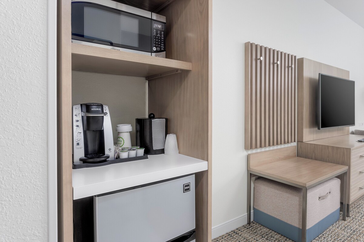 A kitchenette with a microwave, coffee maker, and mini-fridge. There is a TV mounted on the wall and a small seating area with a table and storage.