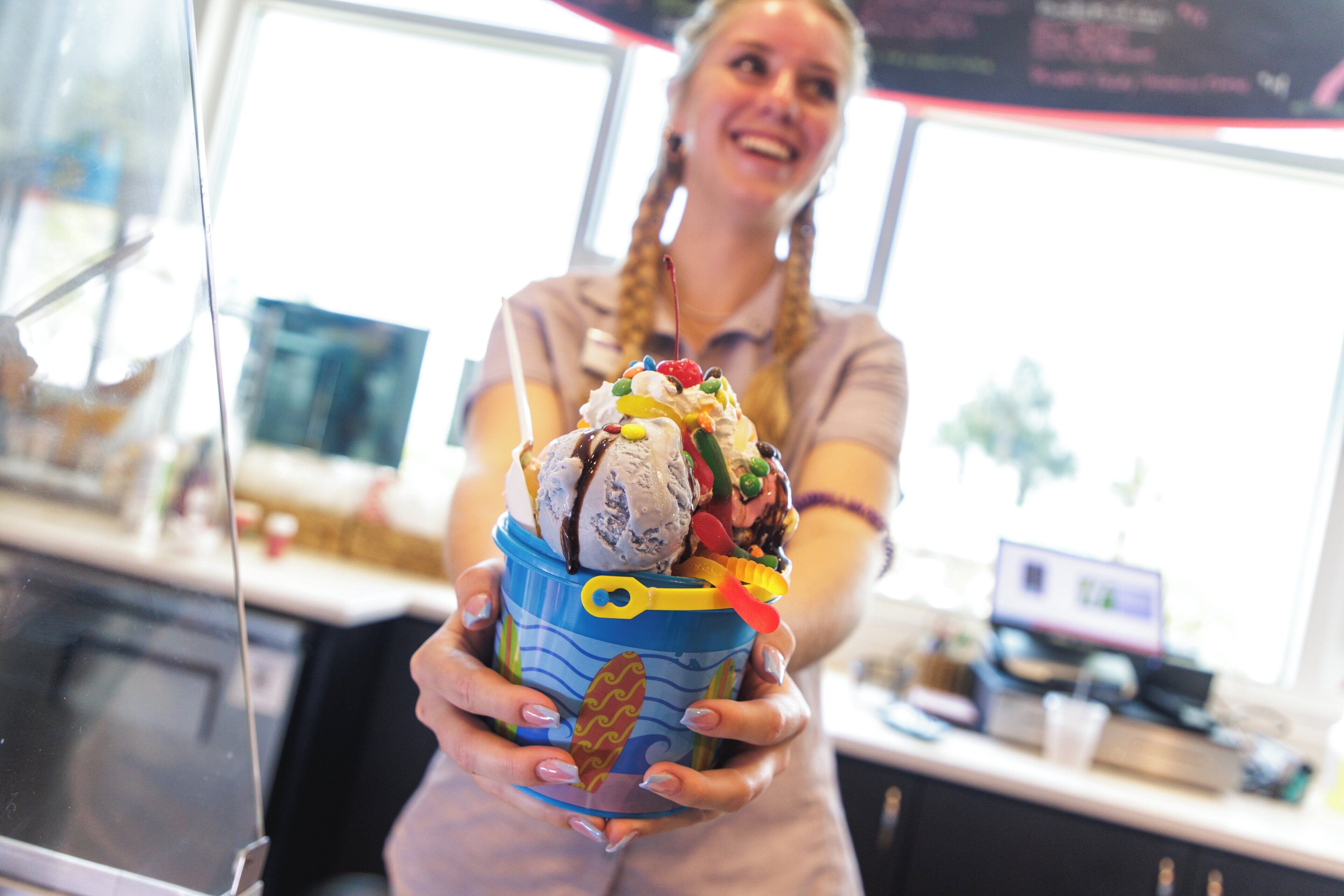 Une personne tient une tasse colorée ornée d’un motif sur le thème de la plage, et cette tasse contient un dessert composé de crème glacée, de fruits et d’une cerise sur le dessus.