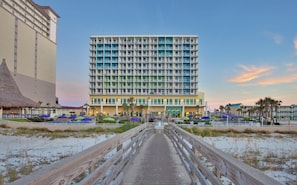 A beachfront hotel with a central courtyard, a boardwalk leading to the entrance, and a thatched-roof structure to the left.