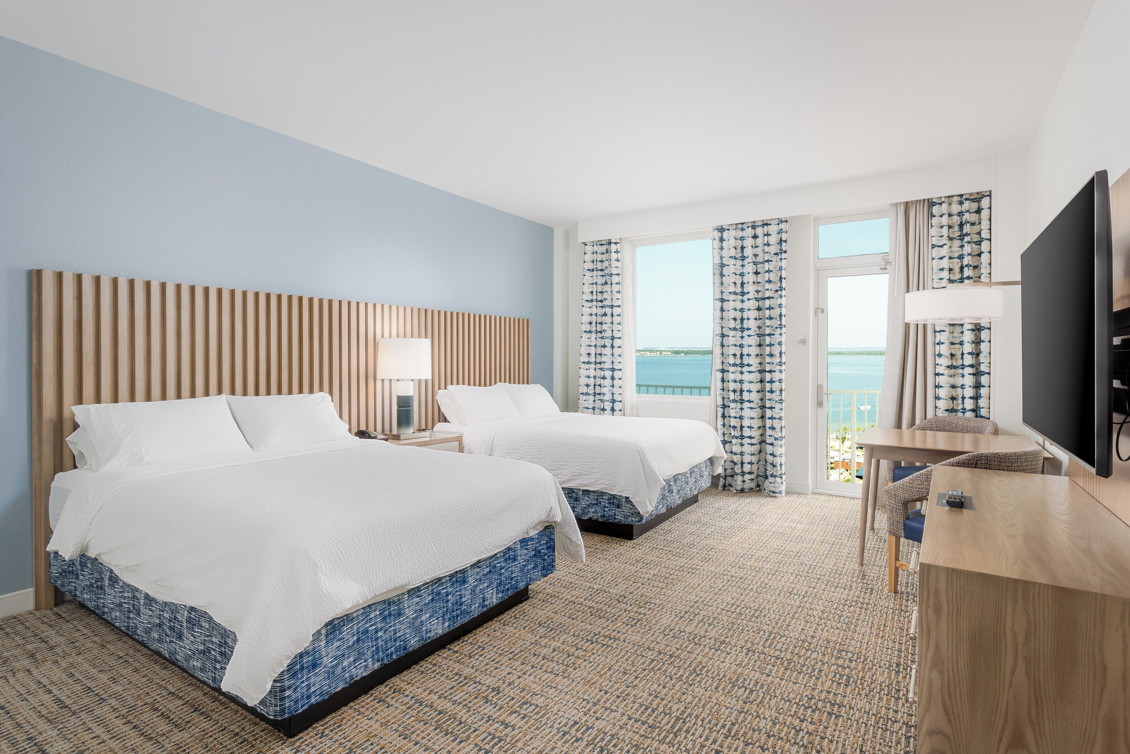 A hotel room with two beds, a desk, a TV, and a view of the sea.