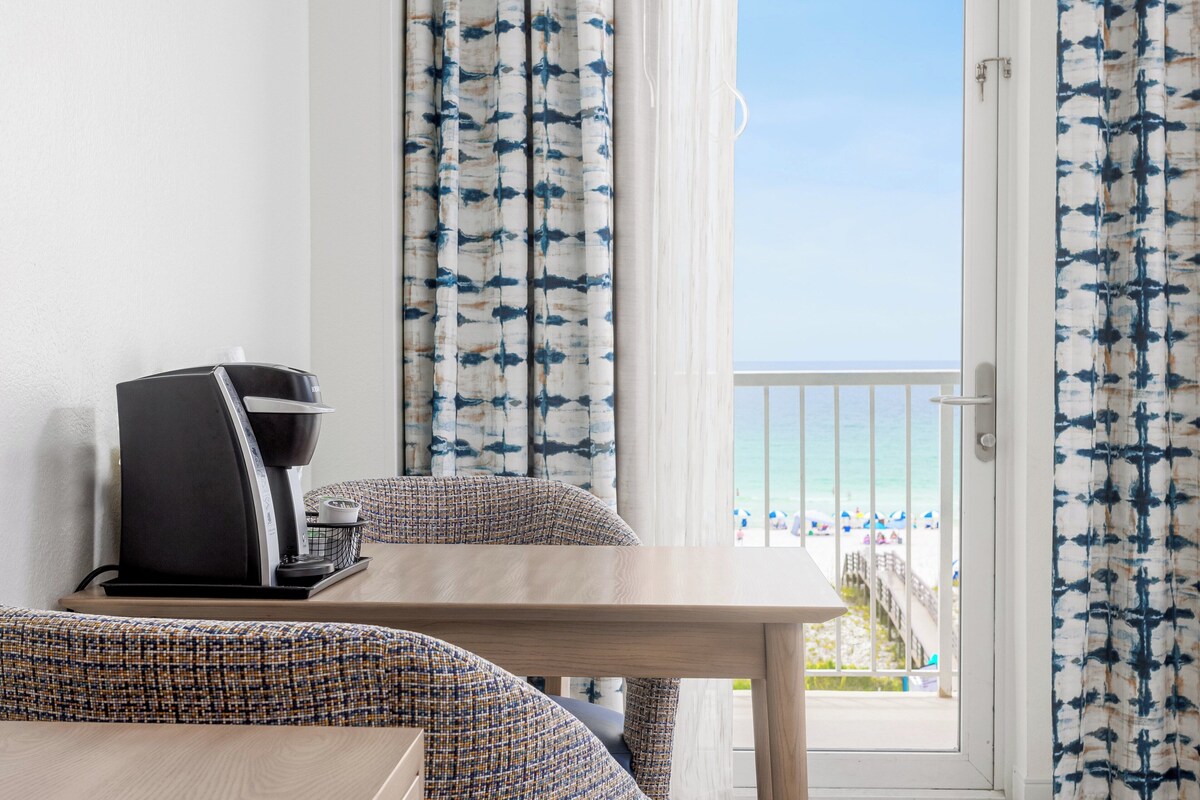 A room with a coffee maker on a table, patterned curtains, and a view of the beach.