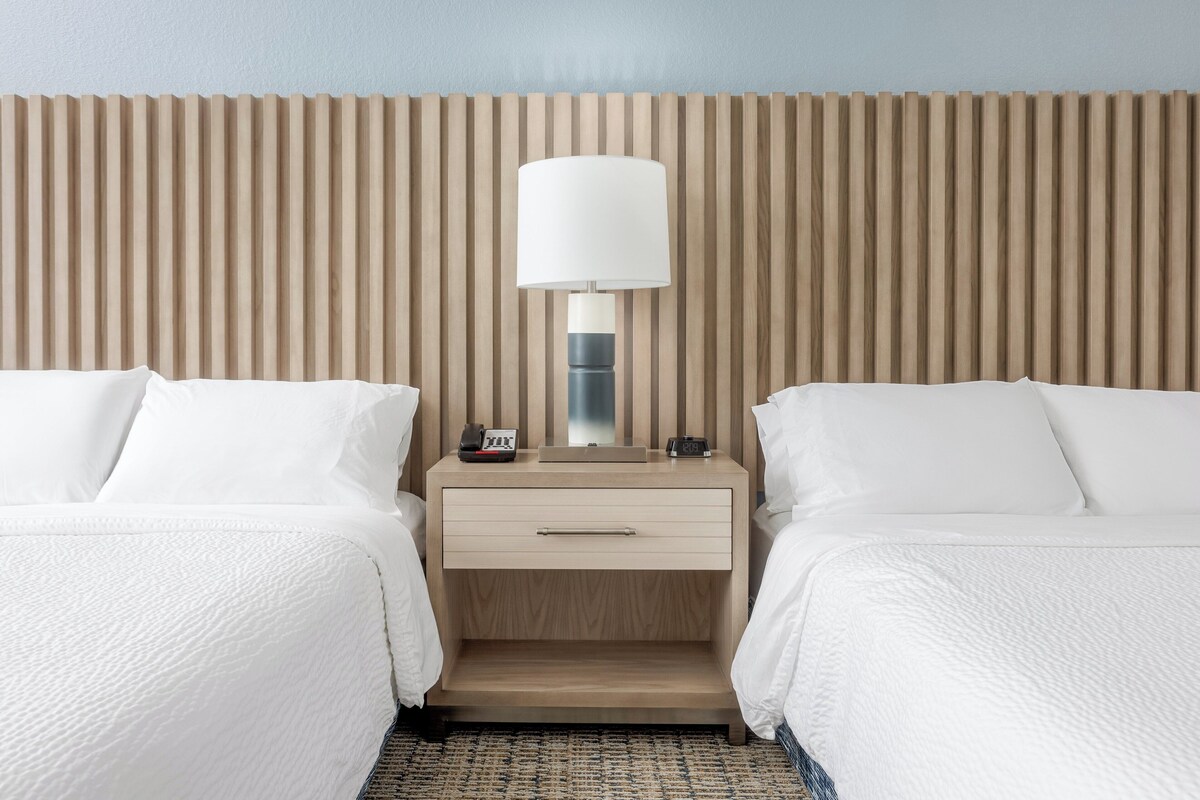 A hotel room with two beds, a nightstand with a lamp, and a wooden slat headboard.