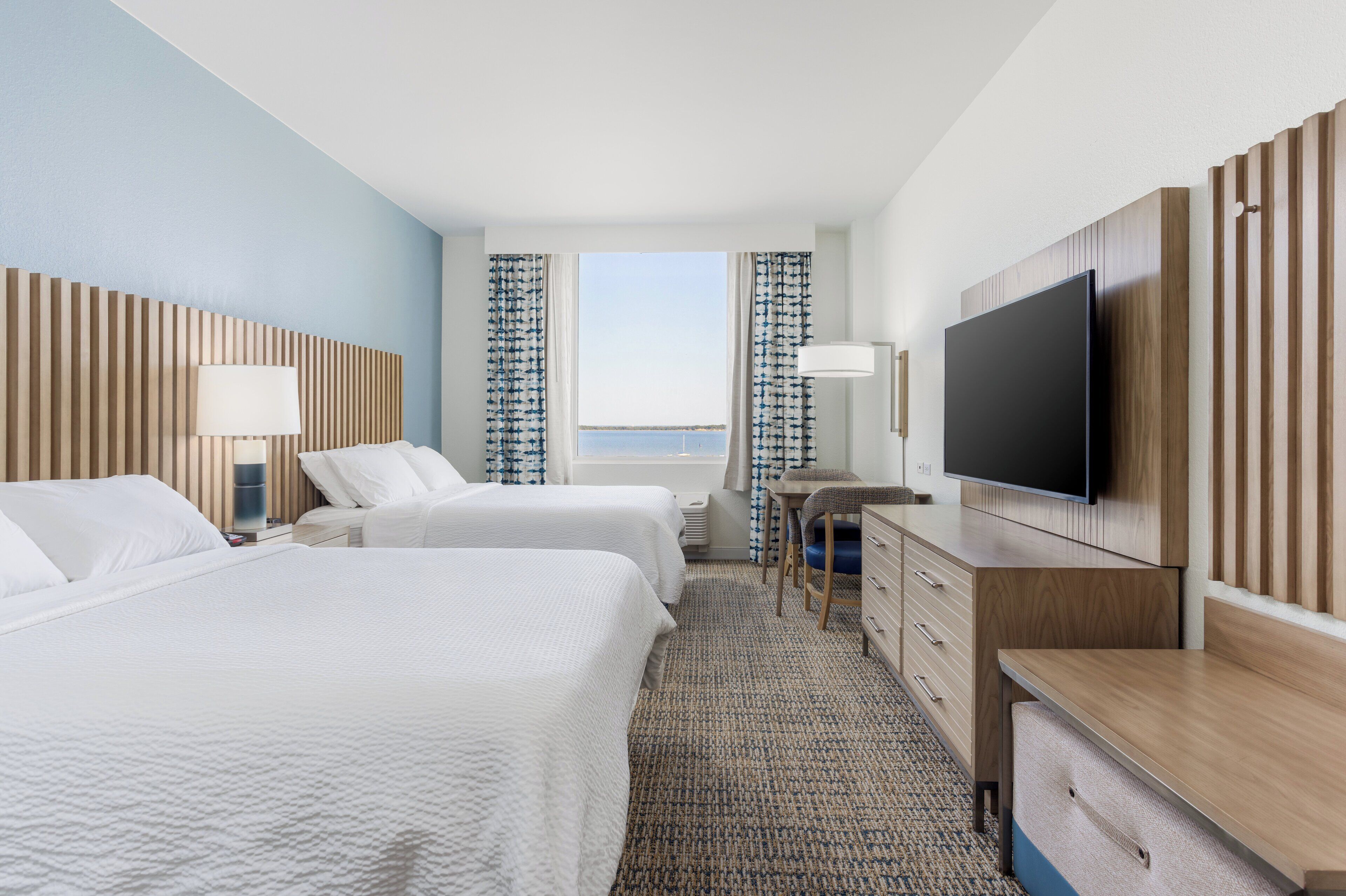 A hotel room with two beds, a desk, a TV, and a view of the sea.