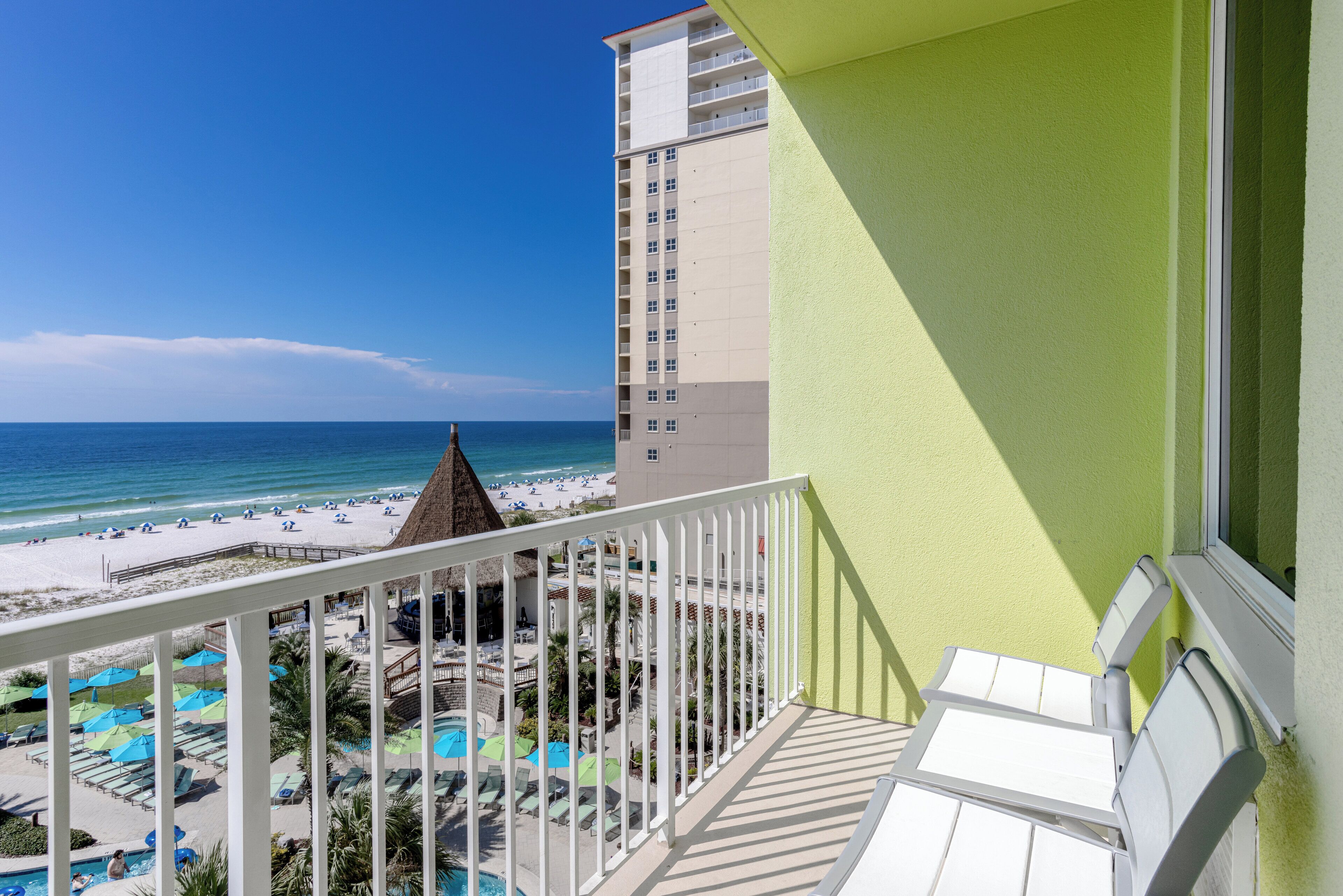 A balcony with a view of a beach, pool area, and a tall building.