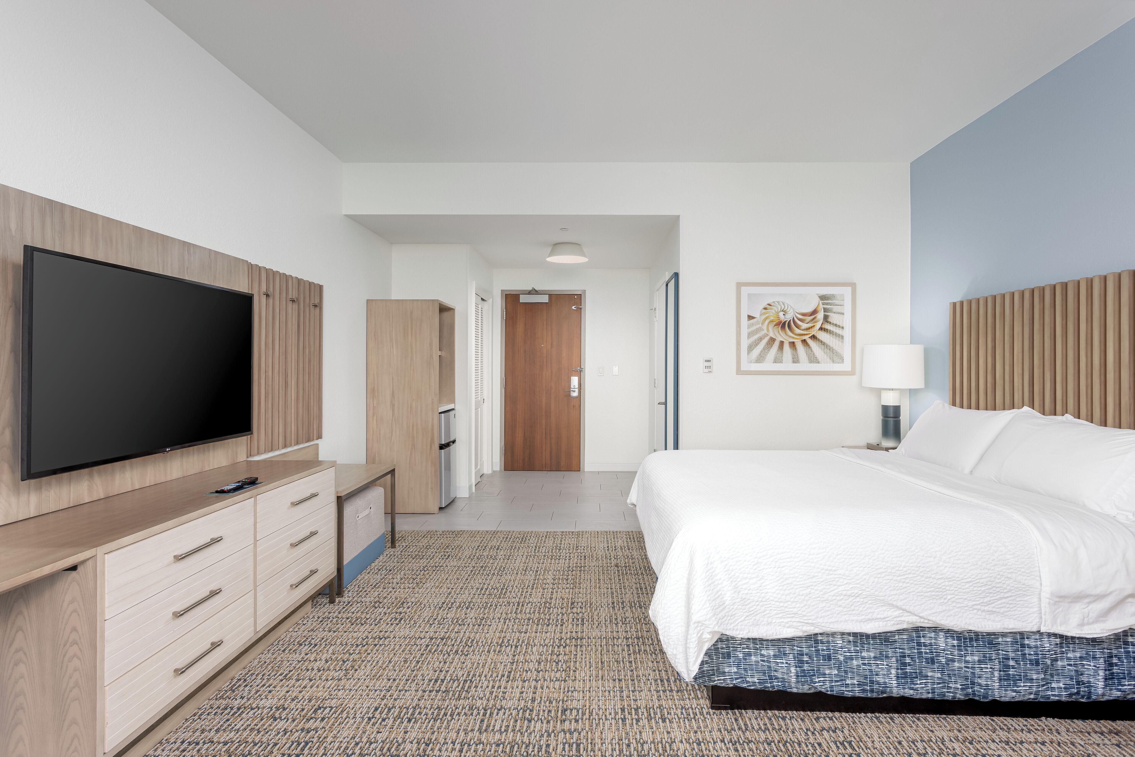 A modern hotel room with a large flat-screen TV, a bed with white bedding, a desk with a lamp, and a patterned rug.
