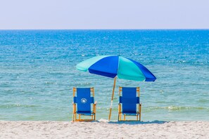 Duas cadeiras de praia sob um guarda-sol azul e turquesa numa praia de areia, com vista para o oceano.
