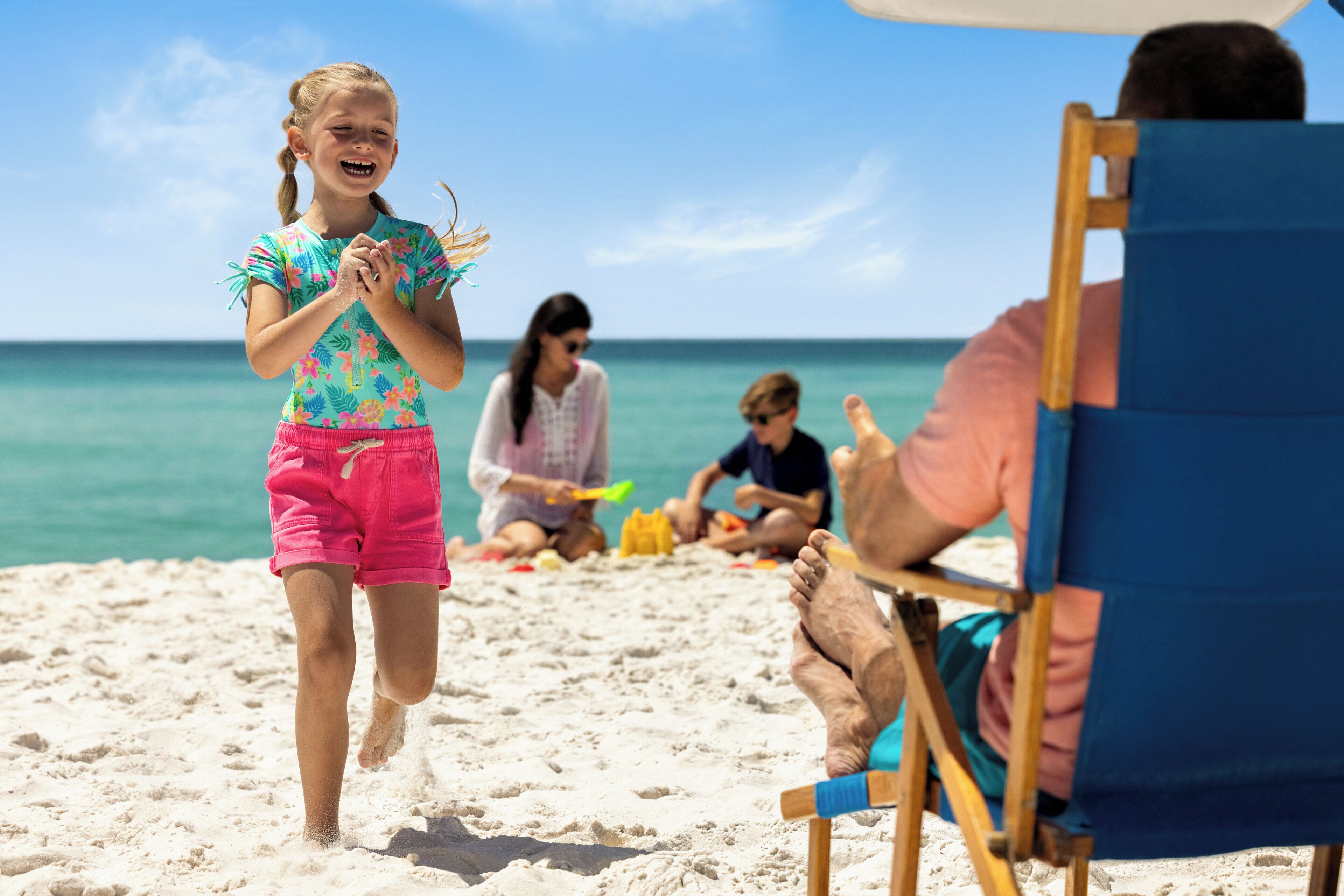 A beach scene with a child running towards the camera, adults sitting on beach chairs, and the ocean in the background.