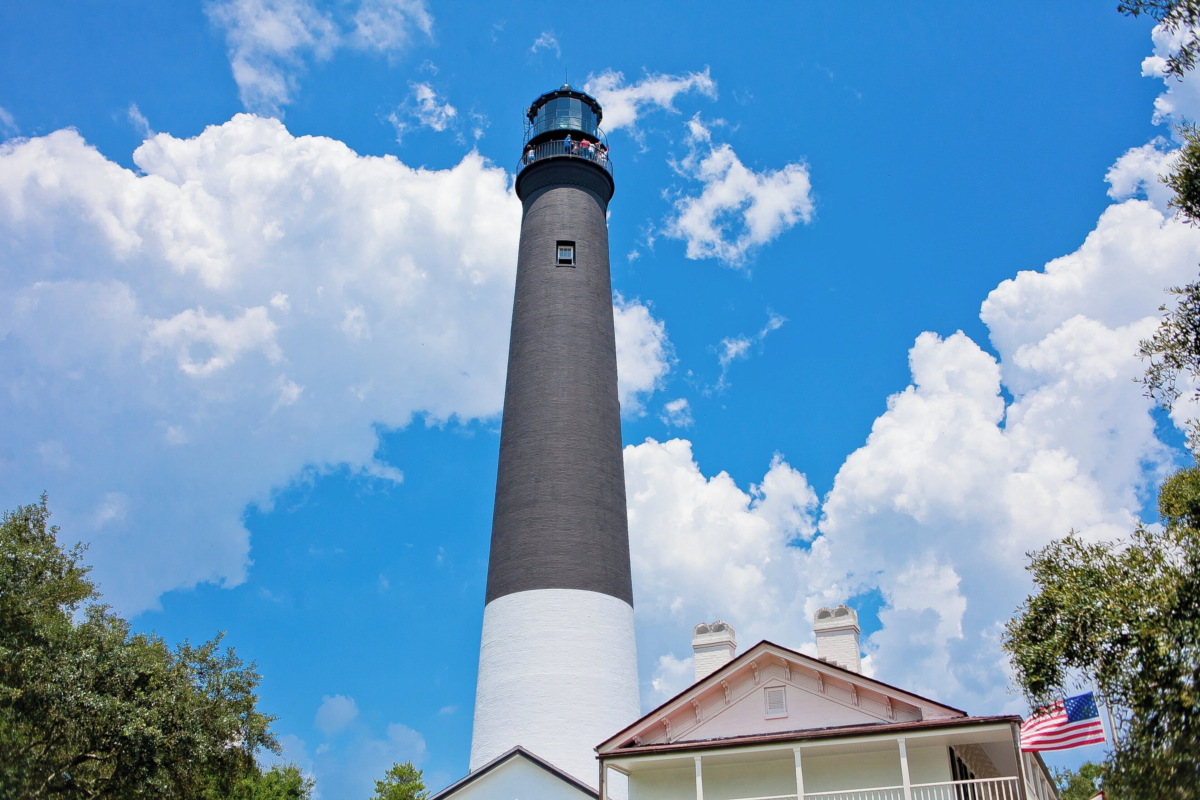 Un grand phare à la base blanche et au sommet foncé, entouré d’arbres et se dressant sous un ciel bleu dégagé.