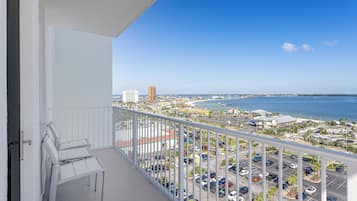 A balcony with white chairs, a view of a parking lot, and a distant beach and ocean.
