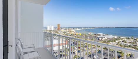 Ein Balkon mit weißen Stühlen, Blick auf einen Parkplatz sowie einen fernen Strand und das Meer.