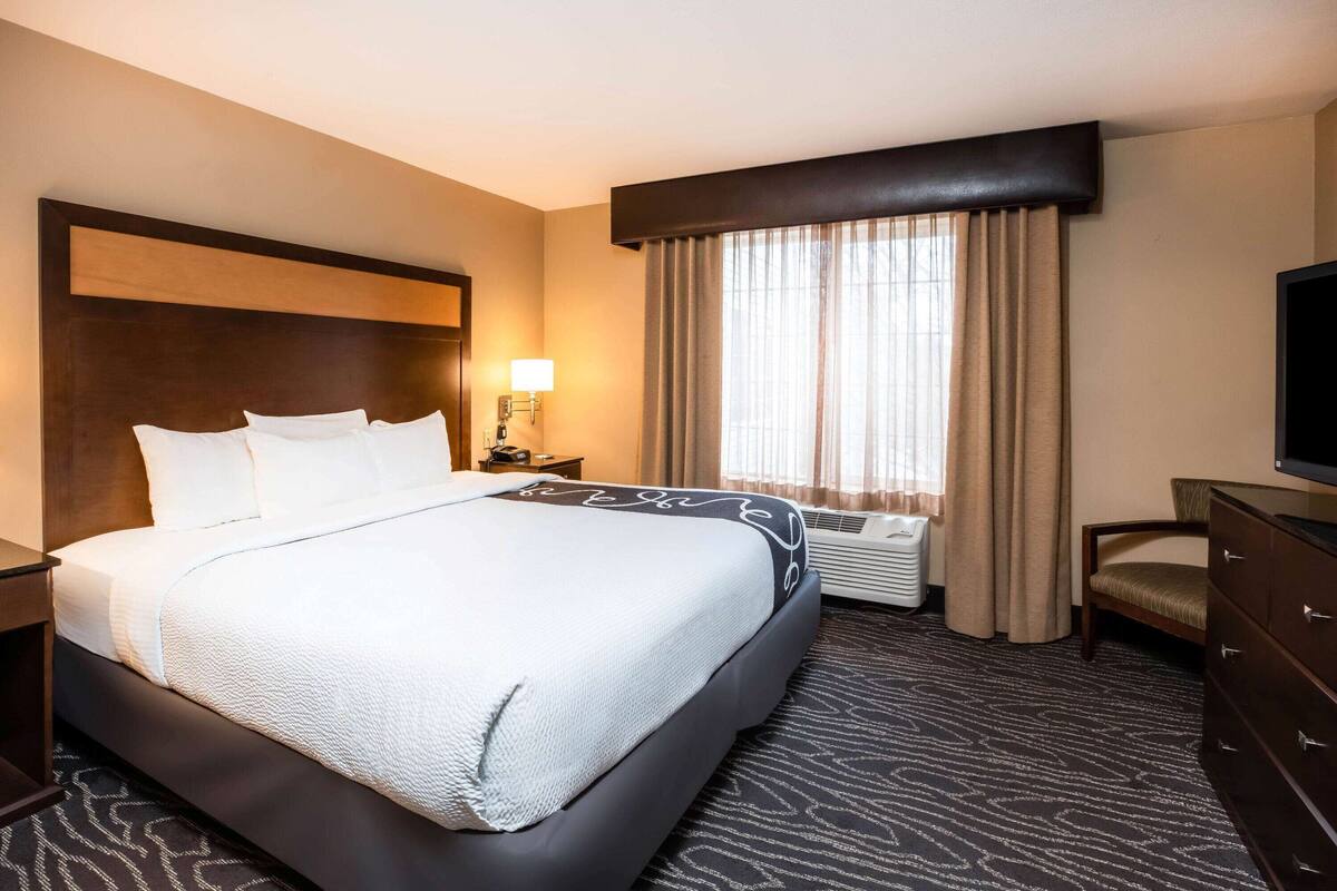 A hotel room with a large bed, a nightstand, a TV, a dresser, and a window with curtains.
