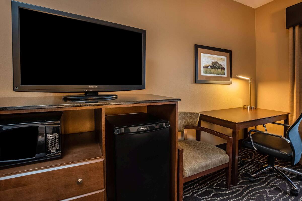 A hotel room with a flat-screen TV, a microwave, a refrigerator, a desk, a chair, and a framed picture on the wall.