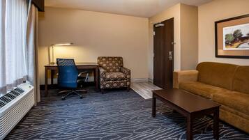 Une chambre d’hôtel avec un bureau, un fauteuil à motifs, un canapé marron, une lampe sur pied et un tableau encadré au mur.