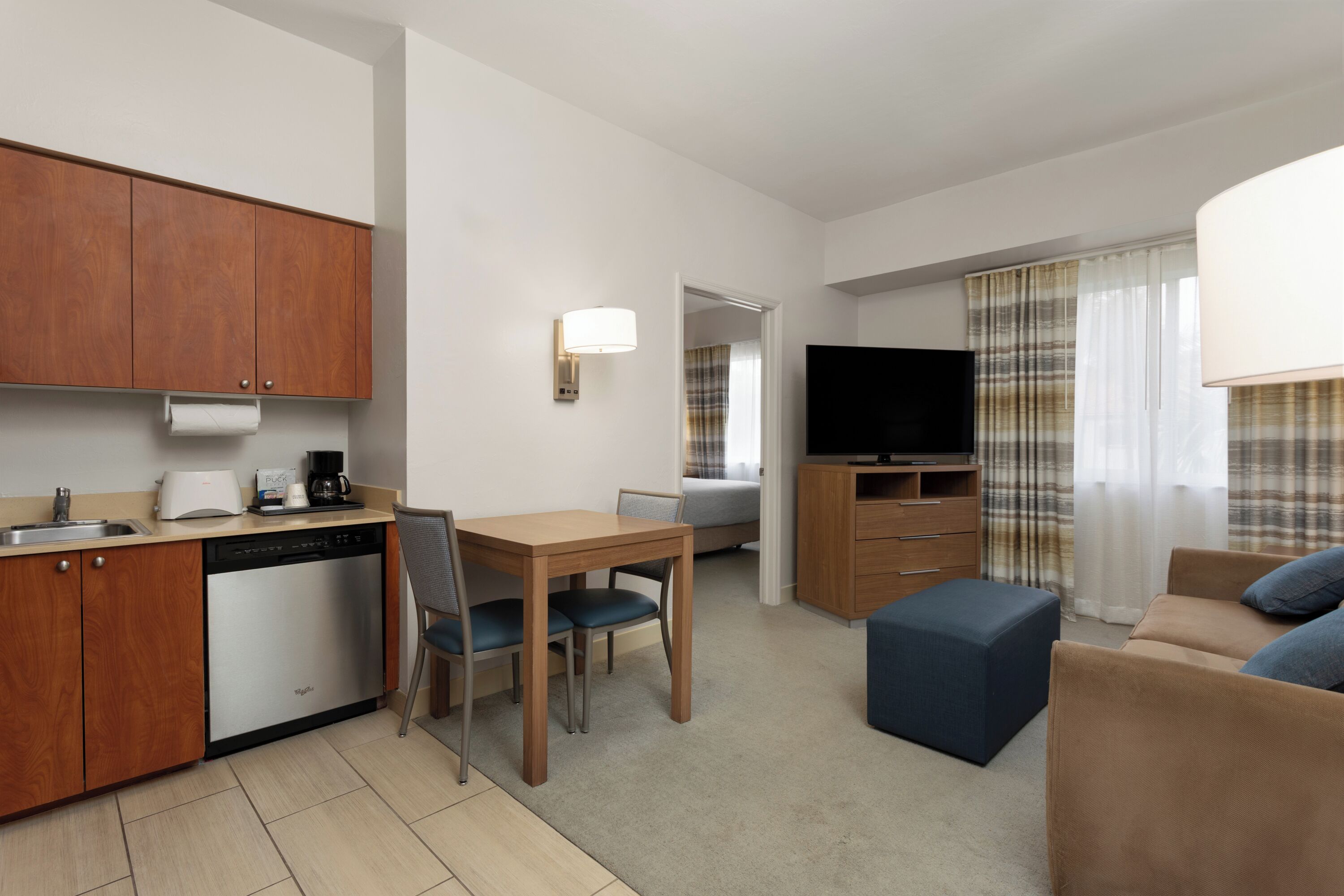 A compact hotel room with a kitchenette, a dining area, a sofa, a TV, and a window with curtains.