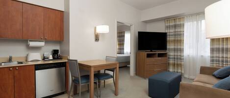 A compact hotel room with a kitchenette, a dining area, a sofa, a TV, and a window with curtains.