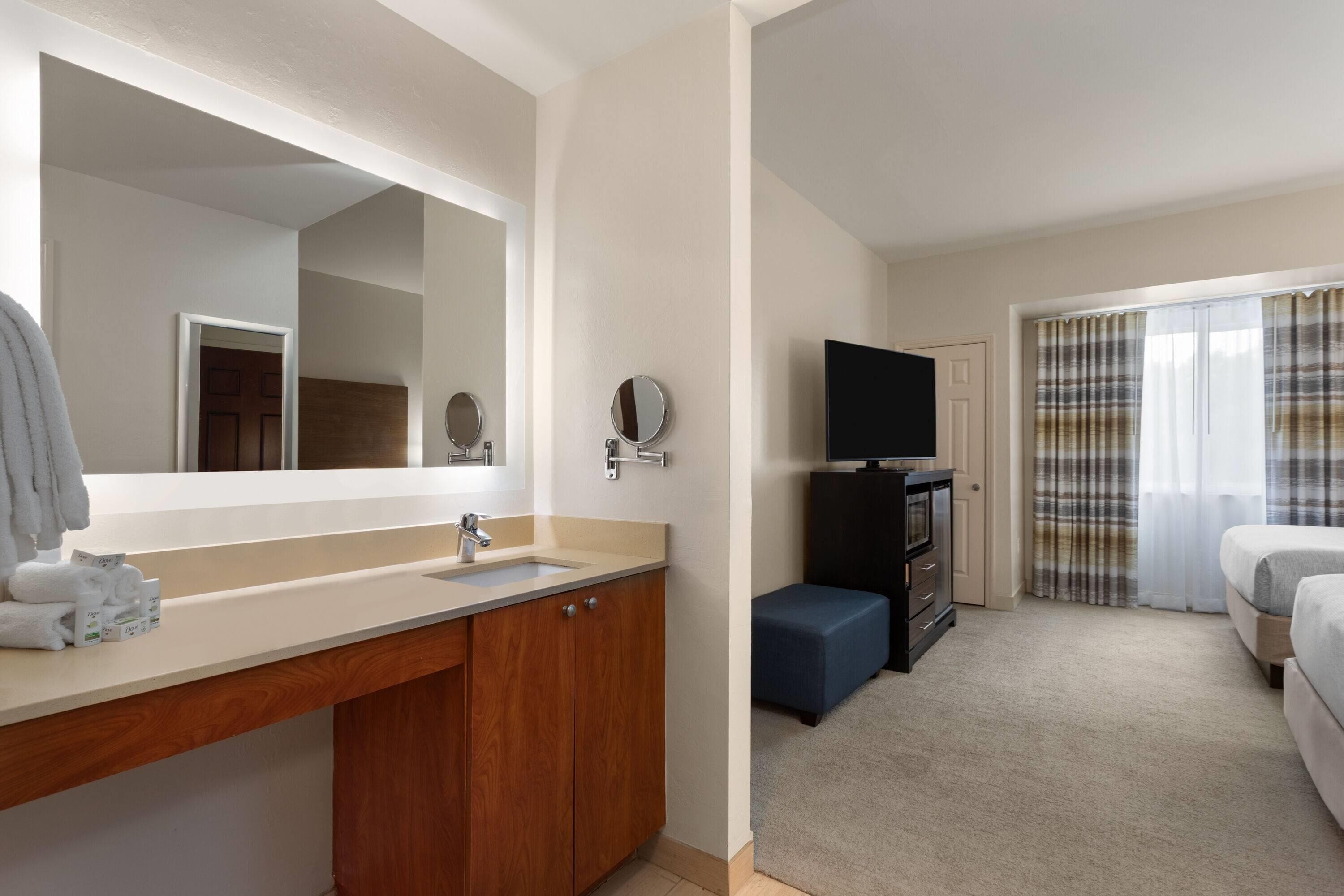 Une chambre d’hôtel avec un grand miroir, un lavabo, une télévision, un lit et une fenêtre avec des rideaux.