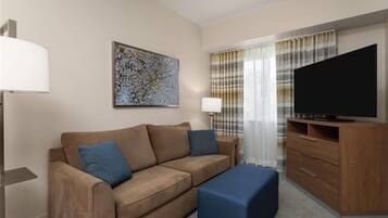 A modern hotel room with a brown sofa, a blue ottoman, a flat-screen TV on a wooden cabinet, a painting on the wall, and a window with curtains.