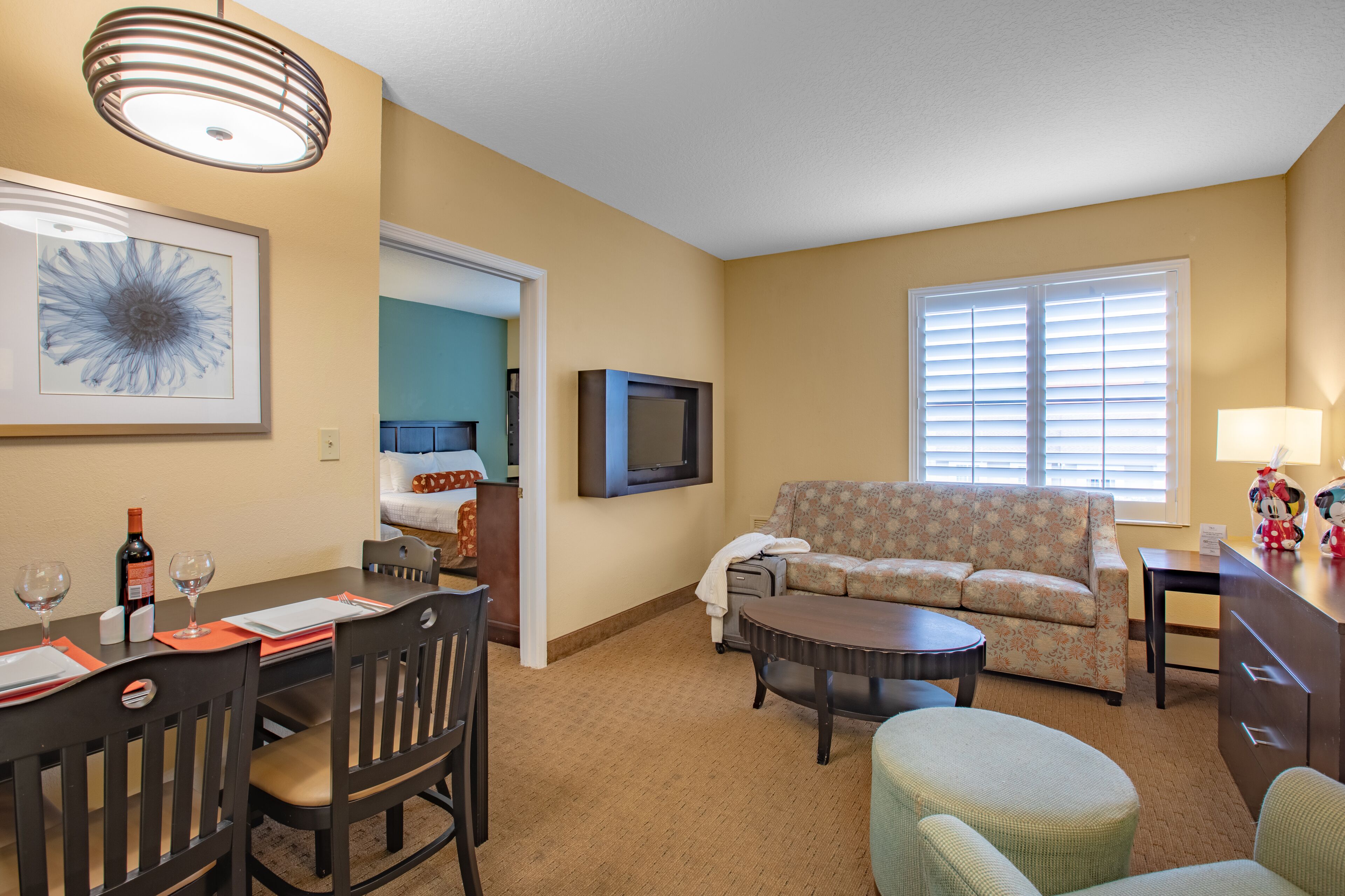 Habitación de hotel con zona de comedor, sofá, televisor y ventana con persianas.