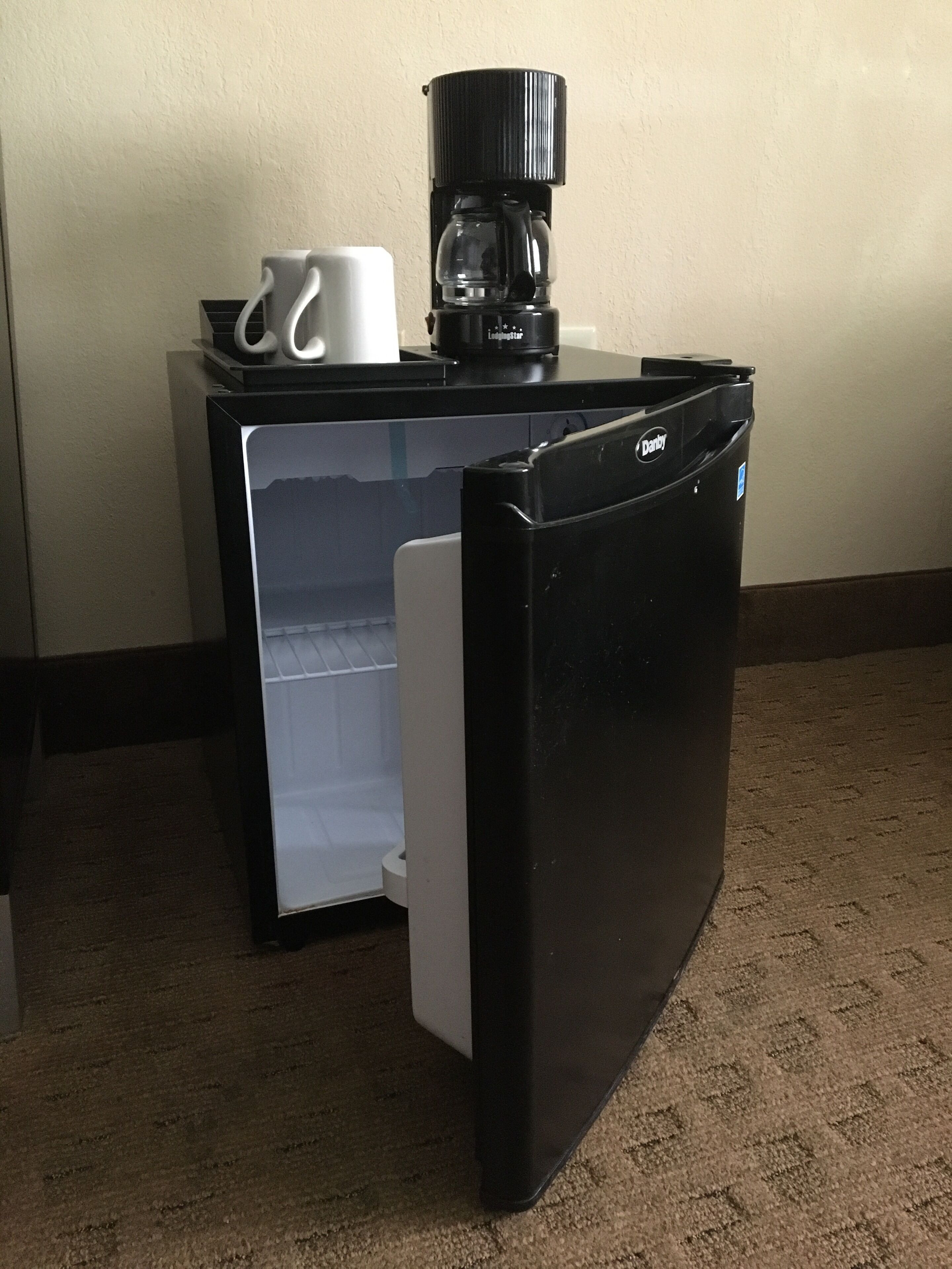 A compact refrigerator with a coffee maker and a mug on top, placed on a carpeted floor.