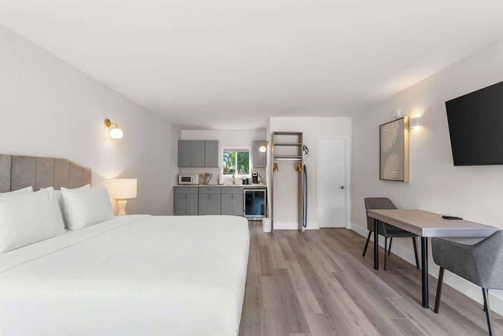 A modern hotel room with a large bed, a desk, and a kitchenette in the background.