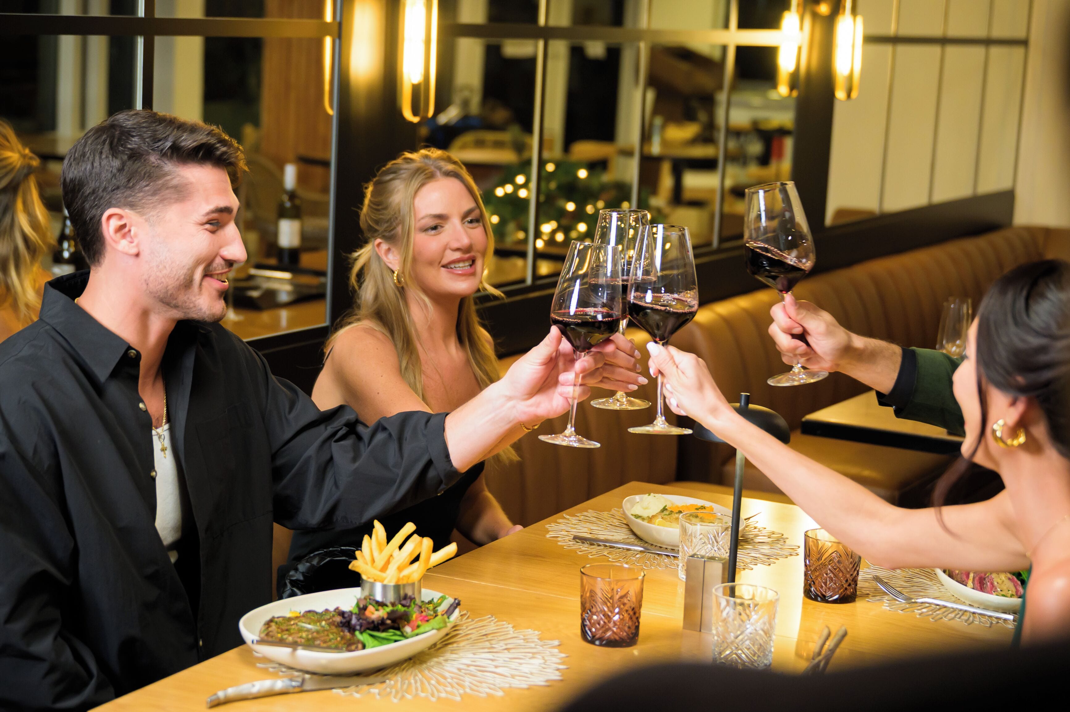 A group of people toasting with wine glasses at a restaurant with a wooden table, plates of food, and a cozy interior.
