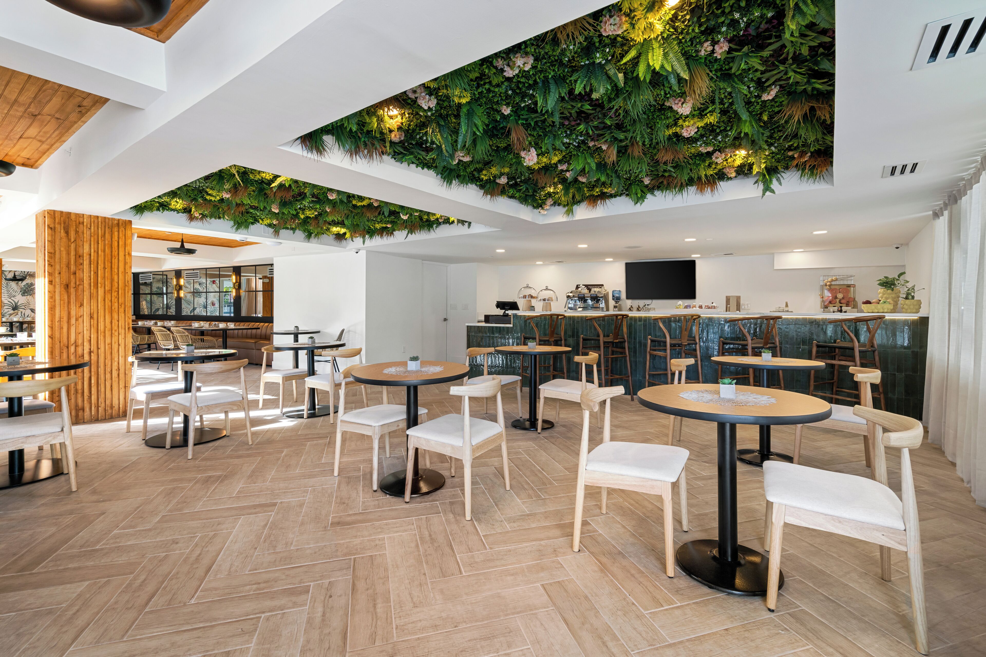 Un café moderno con mesas redondas y sillas blancas, techo de madera y pared verde con plantas.