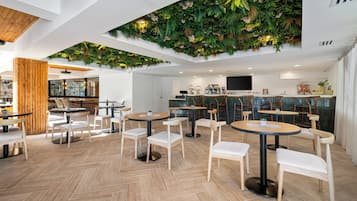 Un café moderno con mesas redondas y sillas blancas, techo de madera y pared verde con plantas.