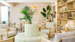 A modern living room with wicker furniture, a central white pouf, and a wooden bookshelf.