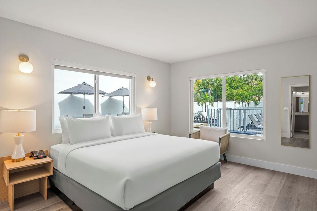 Una habitación de hotel moderna con una cama grande, dos lámparas de noche, una mesita de noche, una silla y un balcón con sombrillas.