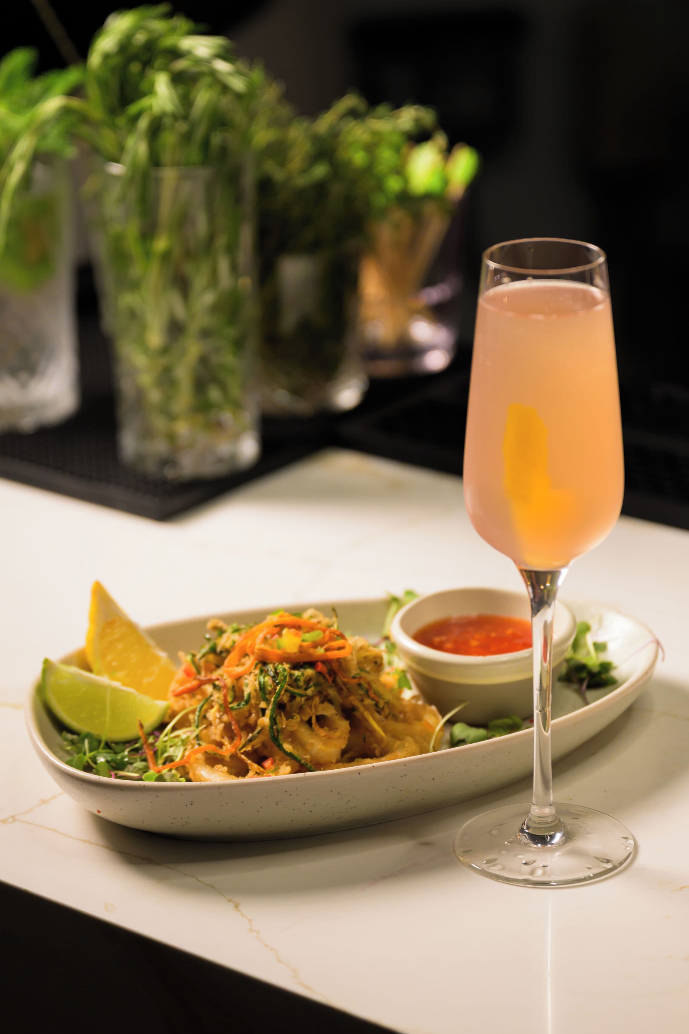 A plate of food with a glass of drink, a lime wedge, and a small bowl of sauce.