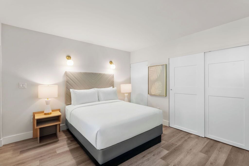 A bedroom with a bed, two bedside lamps, a headboard, a framed artwork, and a wardrobe.